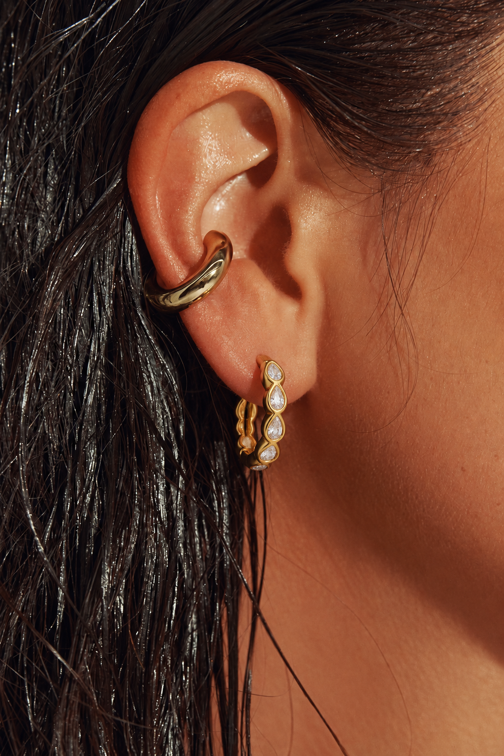 Close-up of a person's ear wearing two gold hoop earrings, including a curved hoop on the upper ear cartilage and a smaller vertical huggie hoop with clear stones on the lobe, against dark hair and warm skin tones.