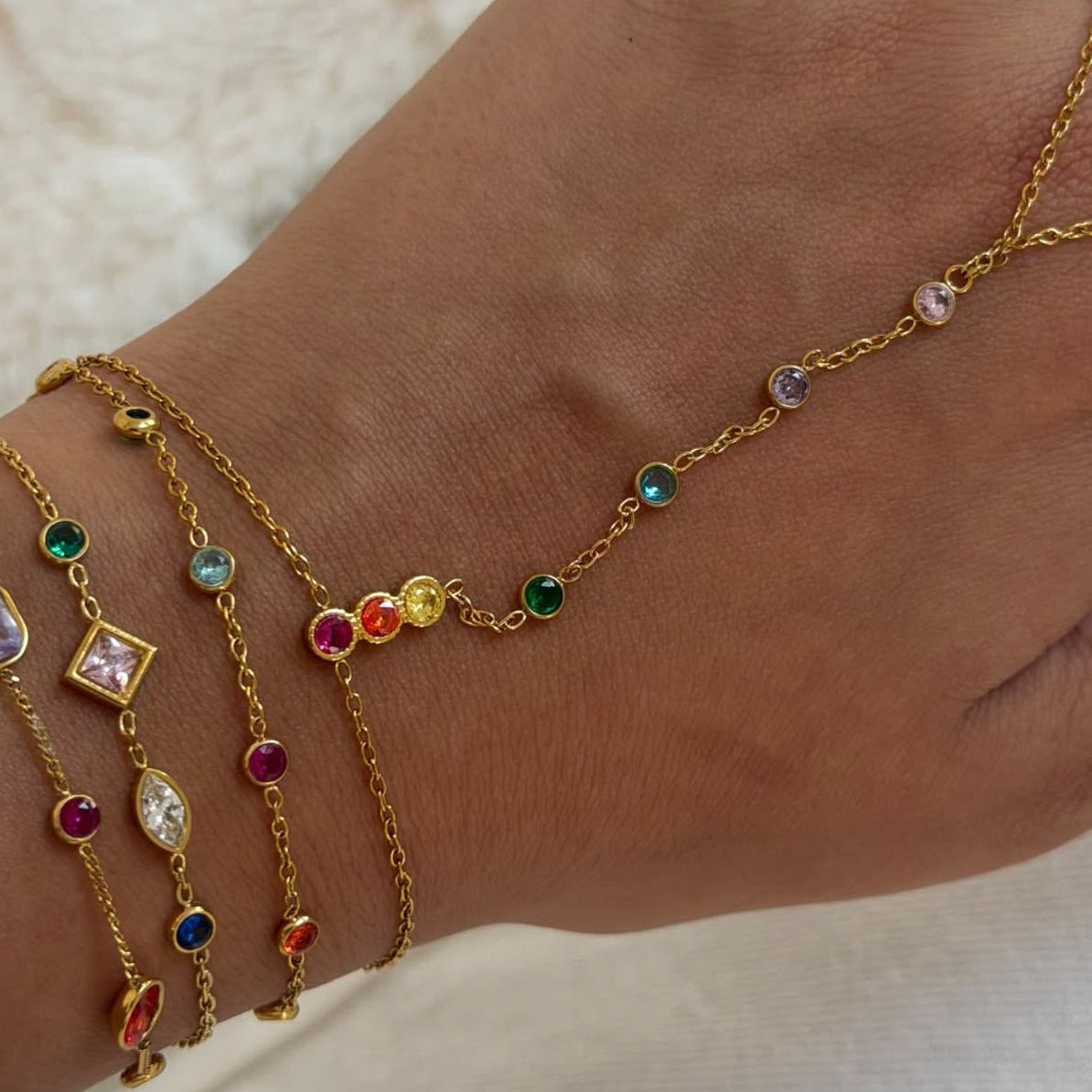 A close-up of a person's ankle and wrist adorned with multiple delicate gold chain anklets decorated with small colorful gemstones, resting on a soft white fabric background.