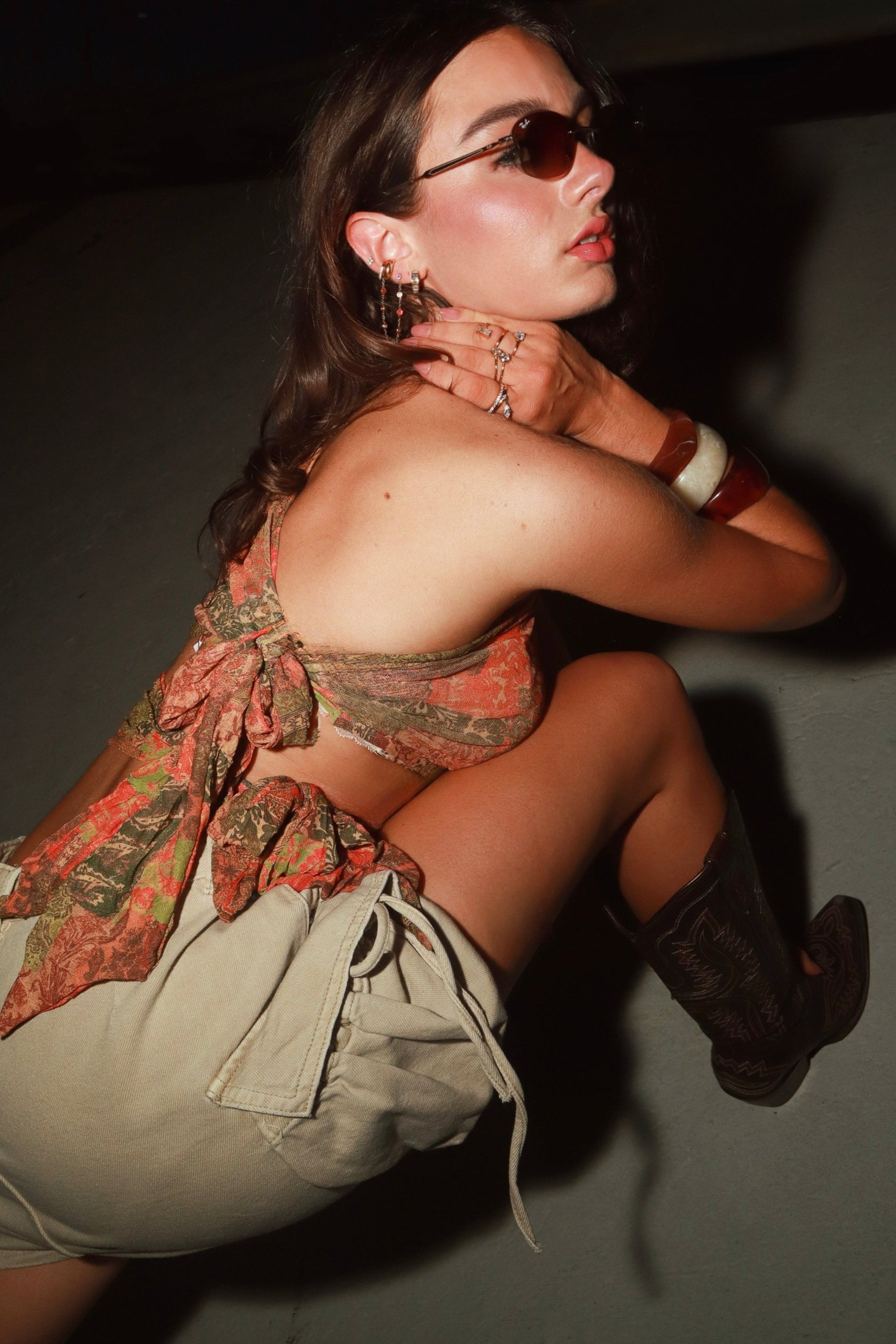 A woman posing in a dimly lit environment wearing a strapless halter top with a rust-orange paisley pattern, light beige cargo shorts, and dark brown cowboy boots.