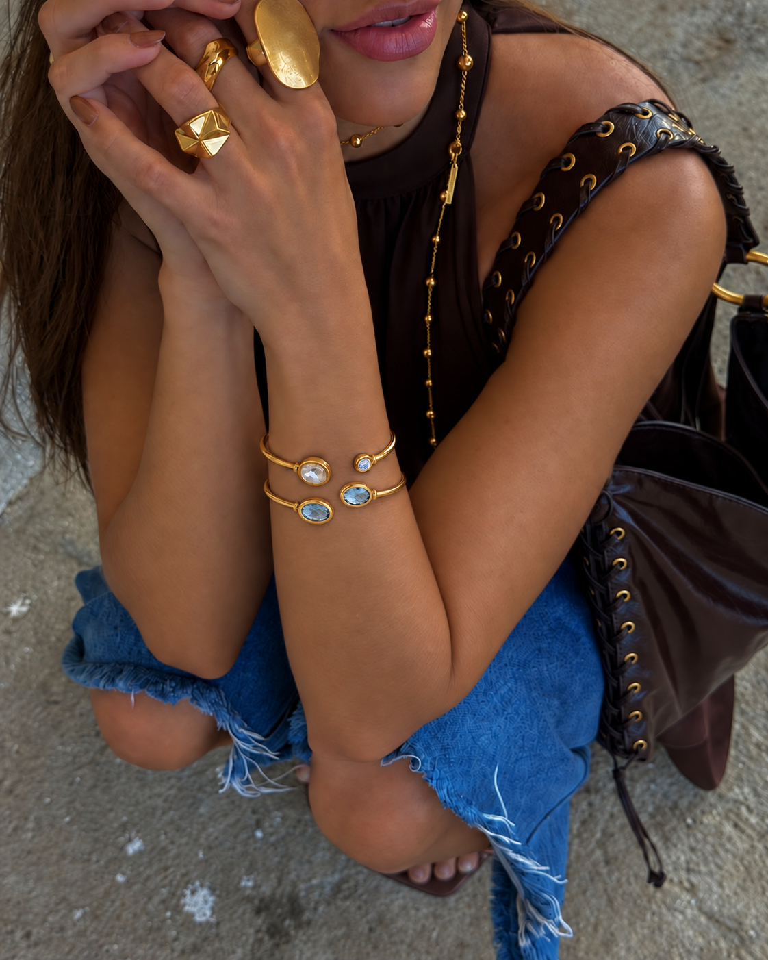 A close-up lifestyle shot of a woman wearing a delicate gold cuff bracelet with two small oval clear stones, styled with gold rings, a gold chain necklace, and a dark brown studded shoulder bag.