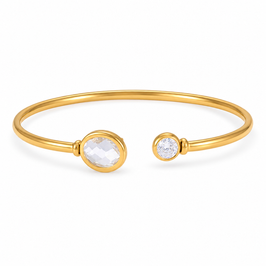 A gold open cuff bangle bracelet featuring a large oval clear faceted gemstone on one end and a smaller round clear gemstone on the other, set against a plain white background.