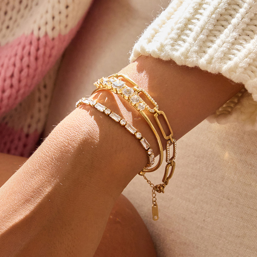 A close-up of a person's wrist wearing three layered gold-tone bracelets, including a chain bracelet with clear rectangular stones and two gold barlink bracelets, styled with a cream knit sweater cuff.