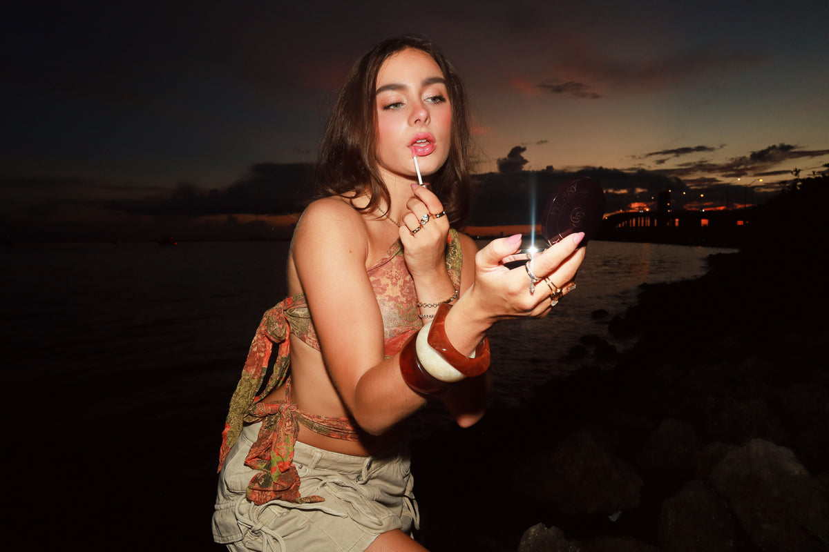 A woman posing on a rocky shoreline at dusk, holding a small lighter and applying lip product. She is wearing a patterned halter top and light-colored shorts, with a dark ocean and cloudy sky in the background.