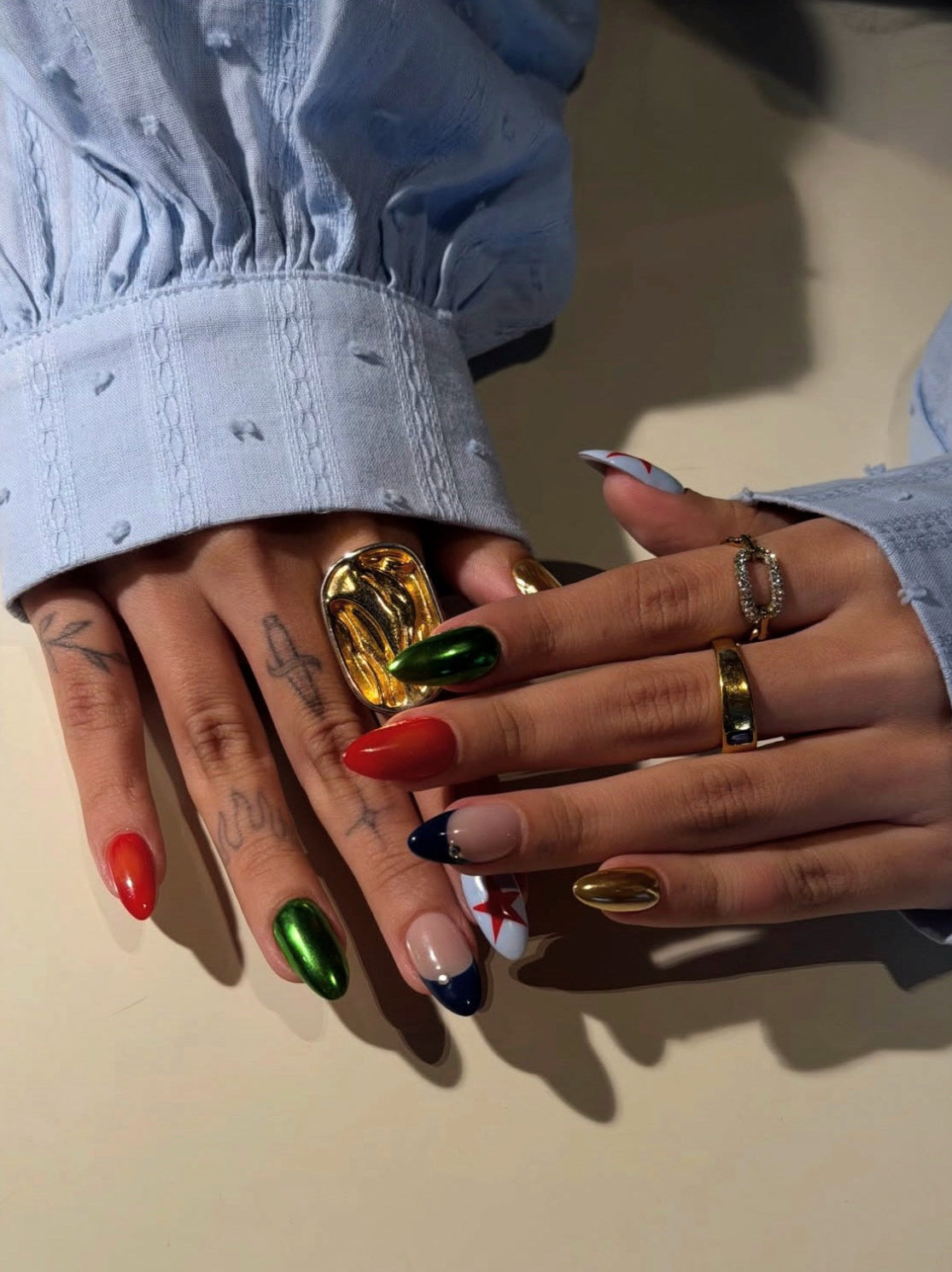 A close-up of two hands with long, manicured nails in red, metallic green, nude, and white with red accents, styled with multiple rings, including a large gold ring. The hands are wearing light blue denim sleeves against a neutral beige background.