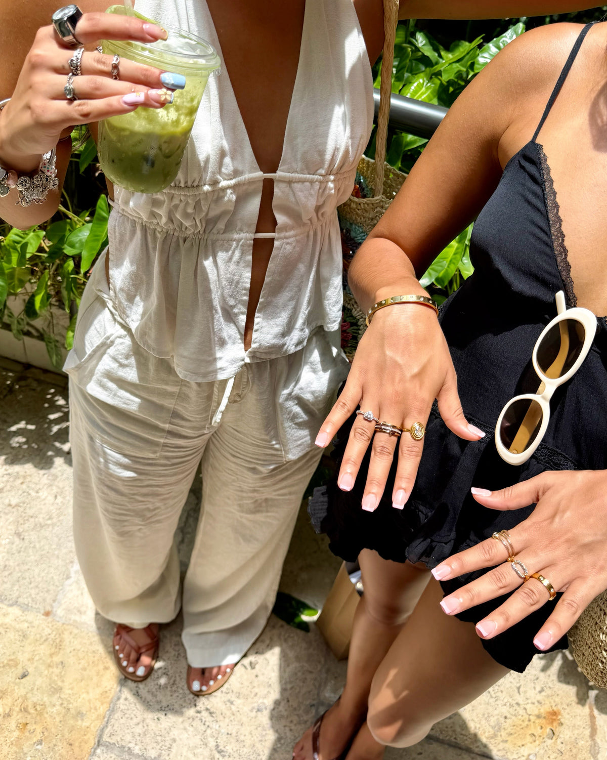 Two women standing outdoors on a stone patio, showcasing summer fashion and accessories. The woman on the left wears a white sleeveless wrap-style jumpsuit and holds a green drink in a glass jar.