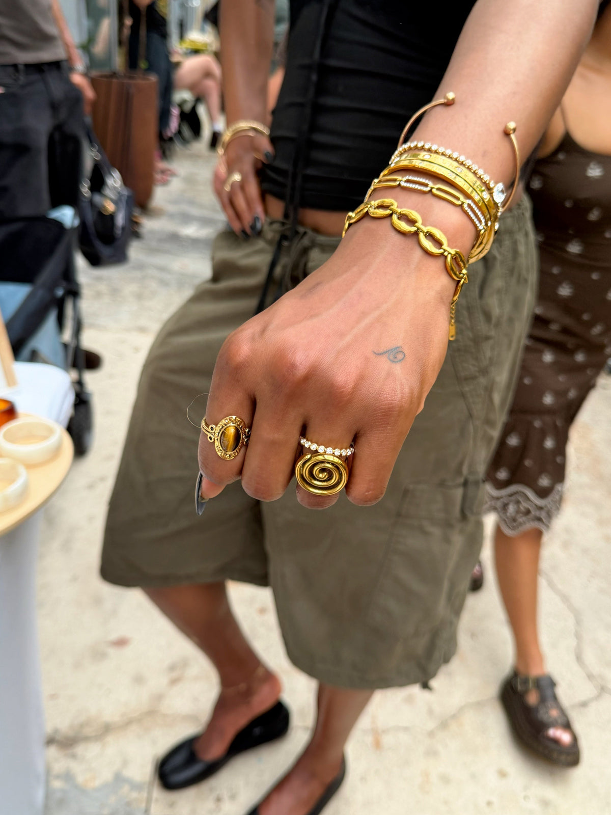 A close-up lifestyle shot of a person's hand showcasing gold jewelry, including a thick chain bracelet, a spiral ring, and stacked bangles, in an outdoor street market setting.