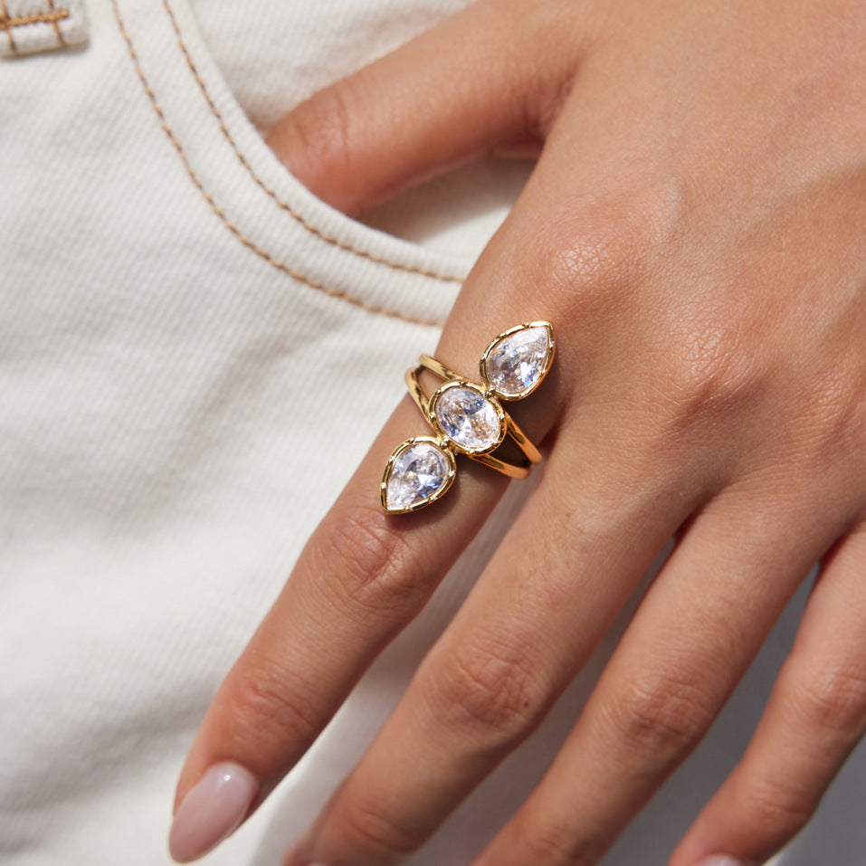A gold ring with three pear-cut clear gemstones in a cluster setting, worn on a person's finger against a white textured fabric background.