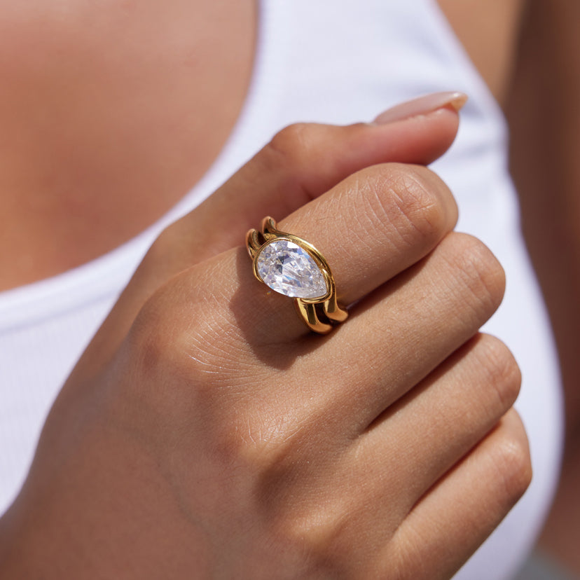 A close-up of a hand wearing a gold ring with a pear-cut clear gemstone, set against a white tank top and blurred skin tones.