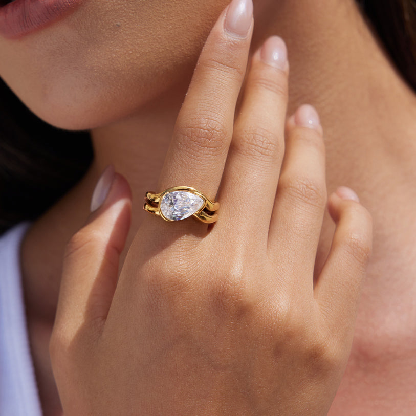 A close-up lifestyle shot of a gold ring with a pear-cut clear gemstone worn on a woman's finger.