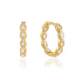 A pair of gold huggie hoop earrings adorned with clear teardrop-shaped crystals, displayed on a clean white background with a soft reflection below.
