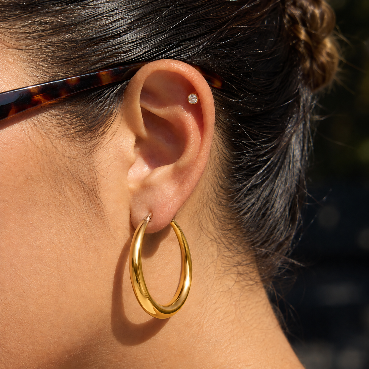 A close-up of a person's ear wearing a polished gold hoop earring and a small diamond stud in the cartilage, with dark hair and part of a tortoiseshell eyeglass frame visible.
