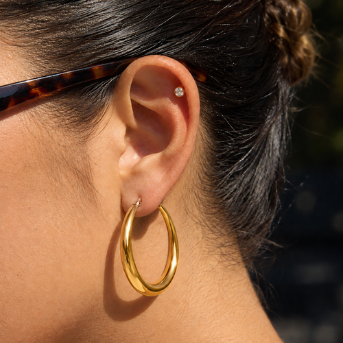 A close-up of a person's ear wearing a polished gold hoop earring and a small diamond stud in the cartilage, with dark hair and part of a tortoiseshell eyeglass frame visible.