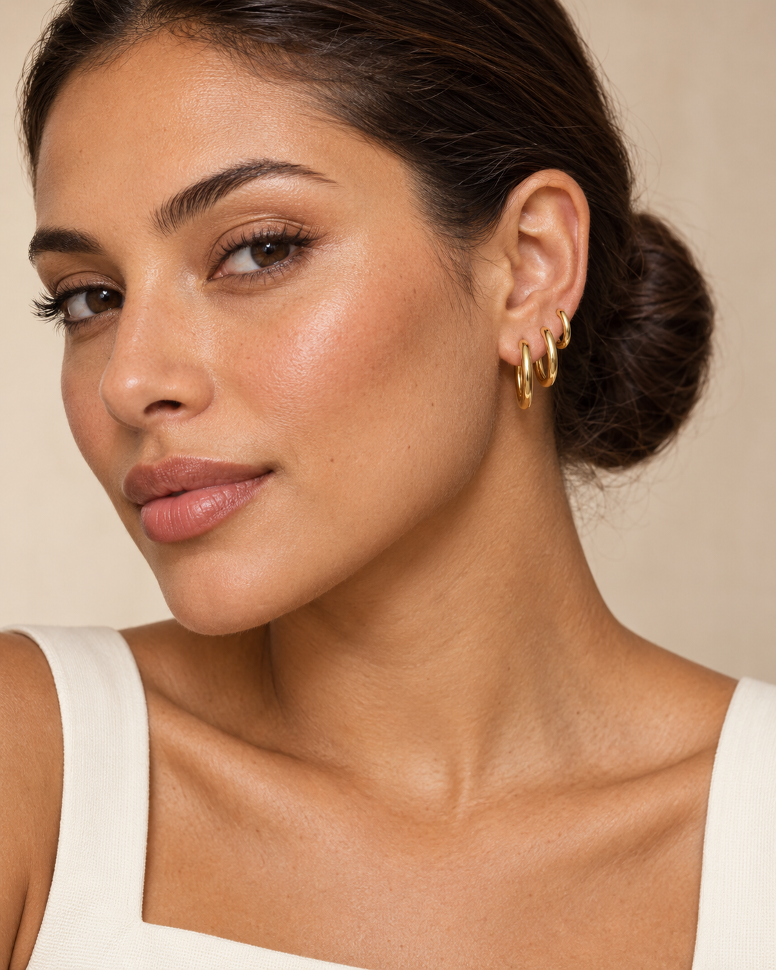 A close-up of a woman wearing three stacked gold hoop earrings, with her hair in a bun and a neutral beige background.