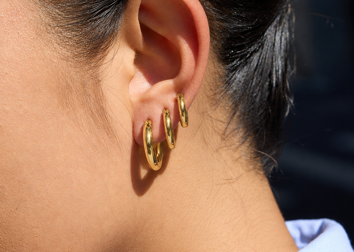 A close-up of a person's ear wearing three small gold hoop earrings stacked on the ear, with dark hair pulled back and a light blue shirt collar visible at the bottom.
